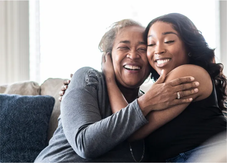 An older woman and a younger woman smile as they hug each other while sitting on a couch.