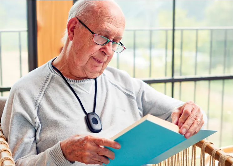 An elderly man wearing a Lively Mobile2 medical alert pendant around his neck looks down at the open book he is holding.